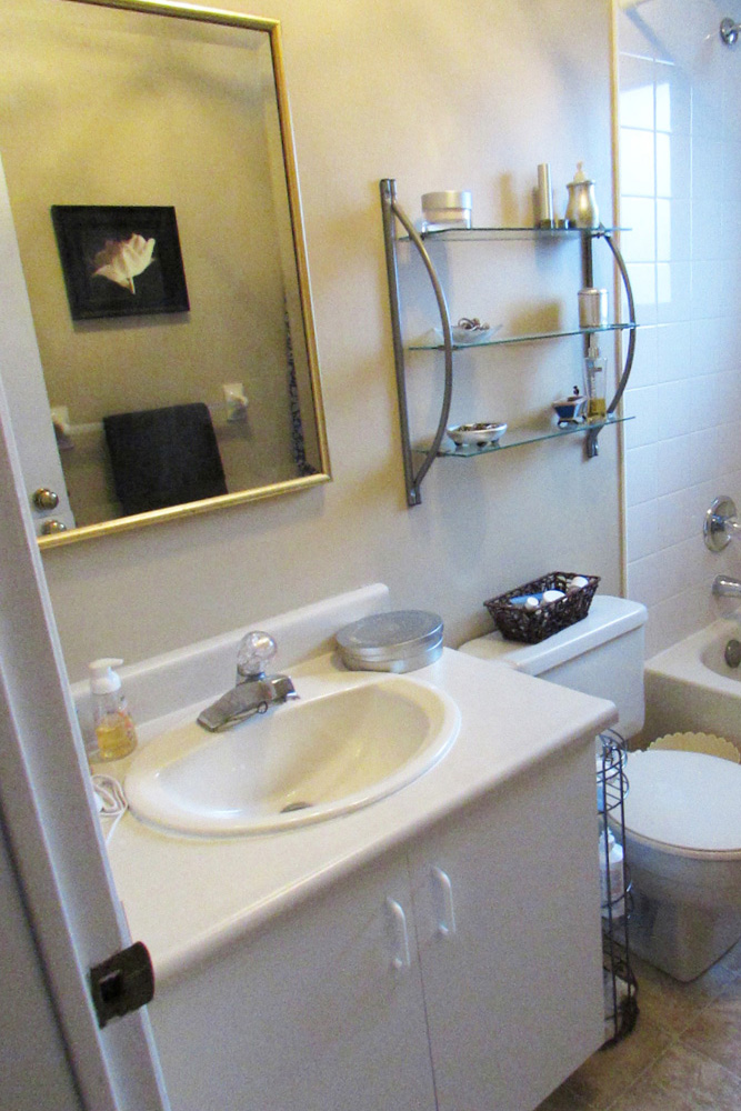 A small bathroom with plain white vanity with a dated faucet, a simple gold mirror and plain glass shelves above the toilet.